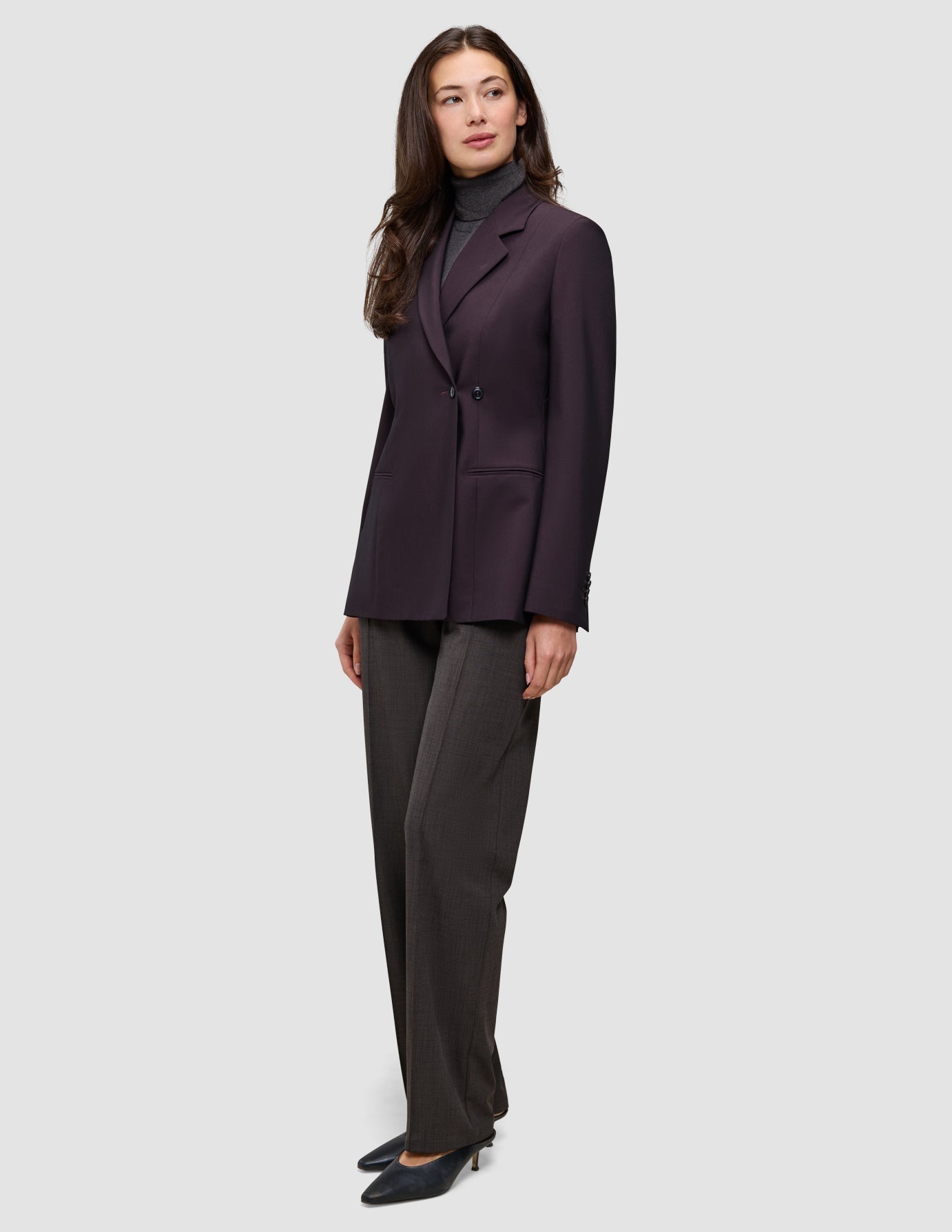 A woman wearing a dark burgundy Women’s Avira Blazer Burgundy Berry double-breasted blazer over a grey turtleneck and grey trousers in a Scandinavian design.